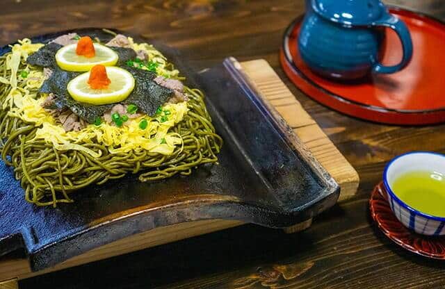 Kawara soba sur une tuile sombre posée sur une planche en bois, avec du thé vert