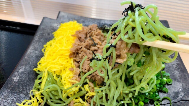 Baguettes soulevant des nouilles soba au thé vert depuis une tuile, à côté d'un œuf et de bœuf