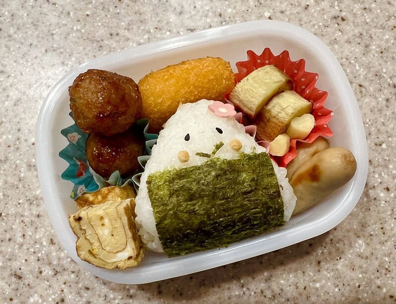 Cute Japanese bento box with rice, onigiri, chicken nuggets, meatballs, tamagoyaki, and pickled vegetables.