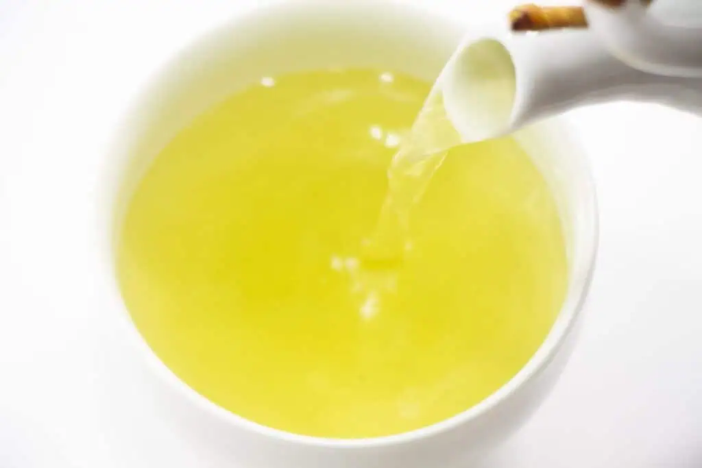 Fresh brewed Asamushi Sencha green tea in a white cup with a teapot pouring.