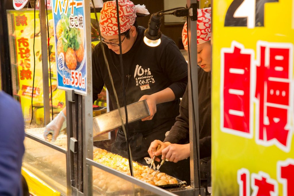 Food vendors preparing Japanese street food at a busy market stall.