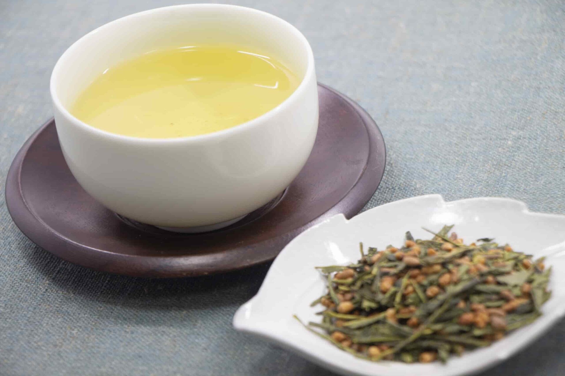 A cup of pale green tea served on a dark wooden saucer, with loose green tea leaves and roasted rice in a white dish beside it.