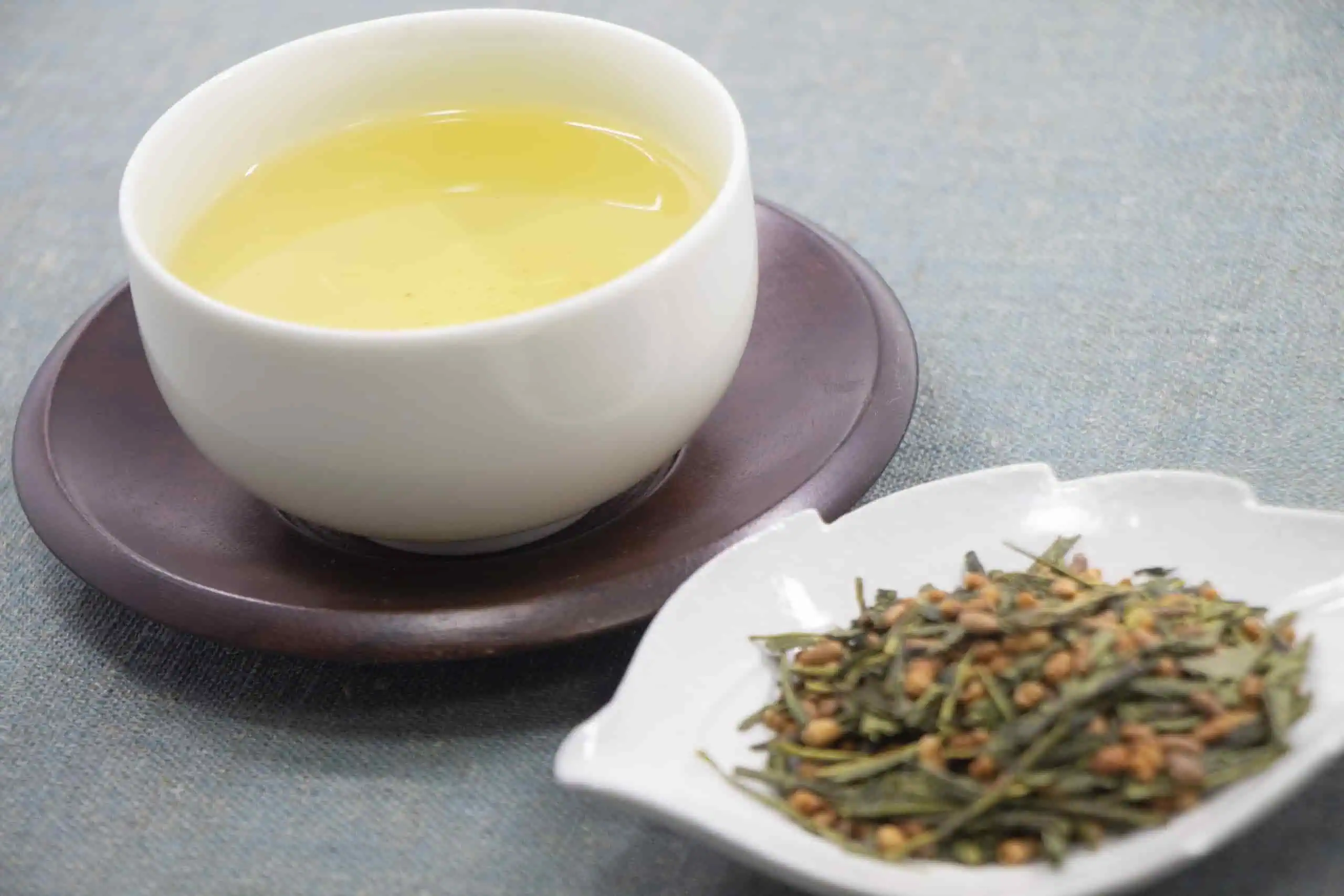 A cup of pale green tea served on a dark wooden saucer, with loose green tea leaves and roasted rice in a white dish beside it.