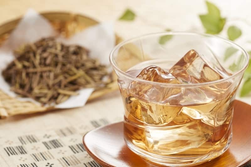 A glass of iced Kaga Bocha with ice cubes, with loose roasted tea stems on paper in the background.