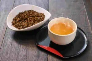 Karigane Hōjicha (雁が音ほうじ茶) A cup of golden-brown karigane hojicha served on a black tray with loose roasted tea stems in a white dish.