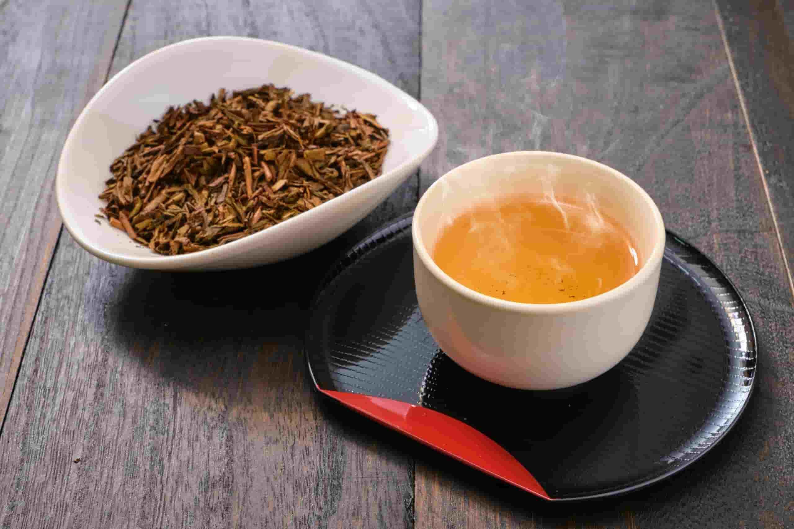 A cup of golden-brown karigane hojicha served on a black tray with loose roasted tea stems in a white dish.