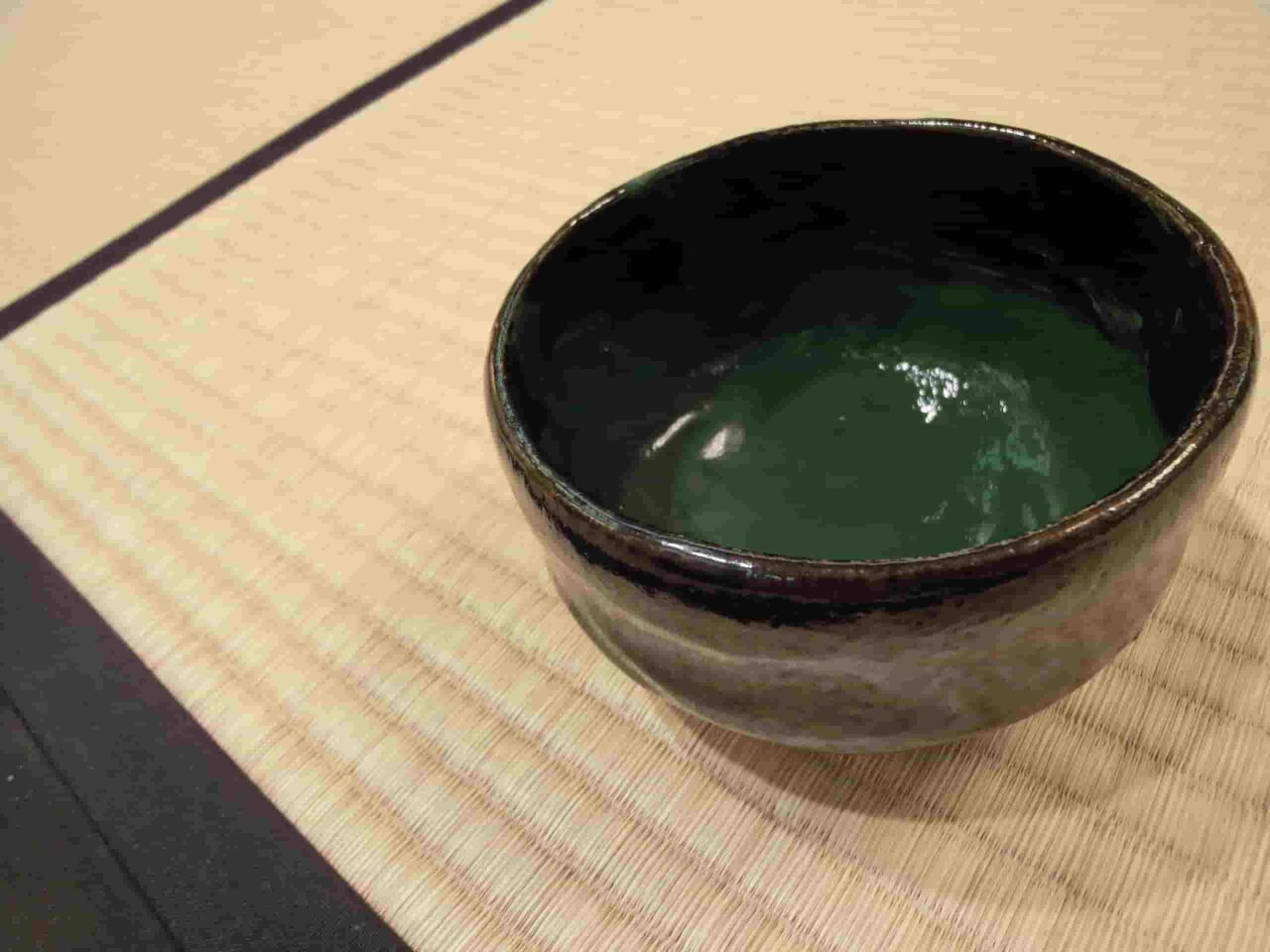 A bowl of thick green koicha tea in a dark ceramic tea bowl on a tatami mat.