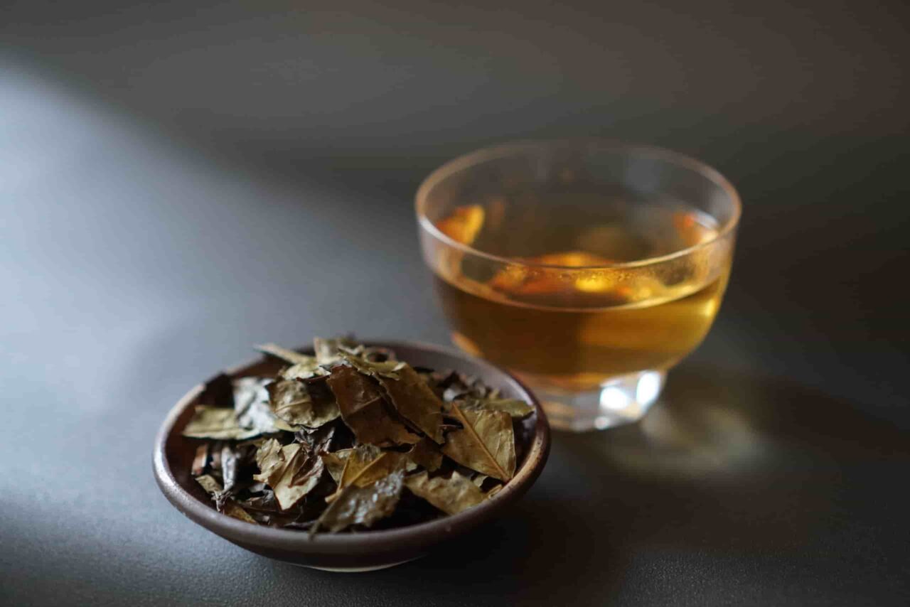 A glass cup of golden-brown tea on a table, with a blurred bowl of dark tea leaves in the background.with leaves