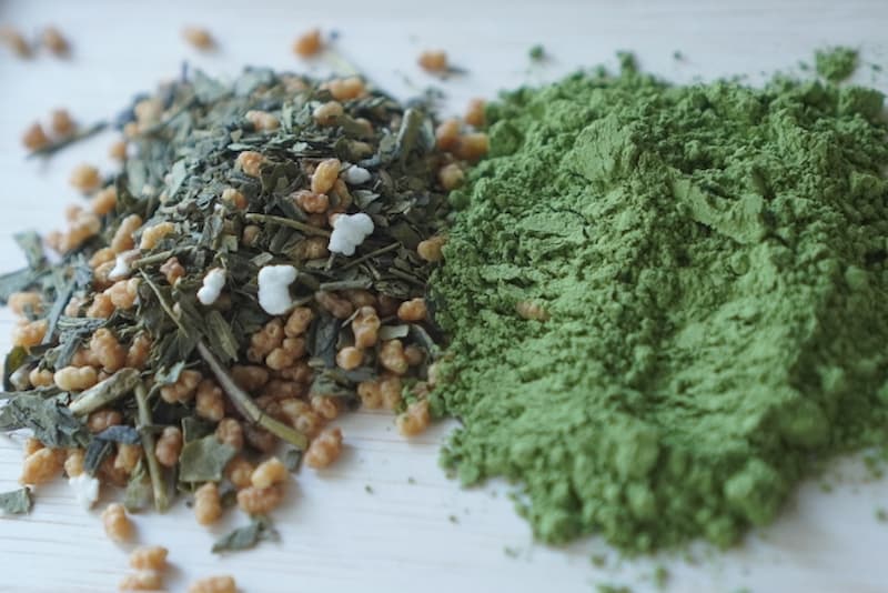 Matcha-iri genmaicha tea leaves with roasted rice and white rice puffs beside a mound of finely ground green matcha powder.