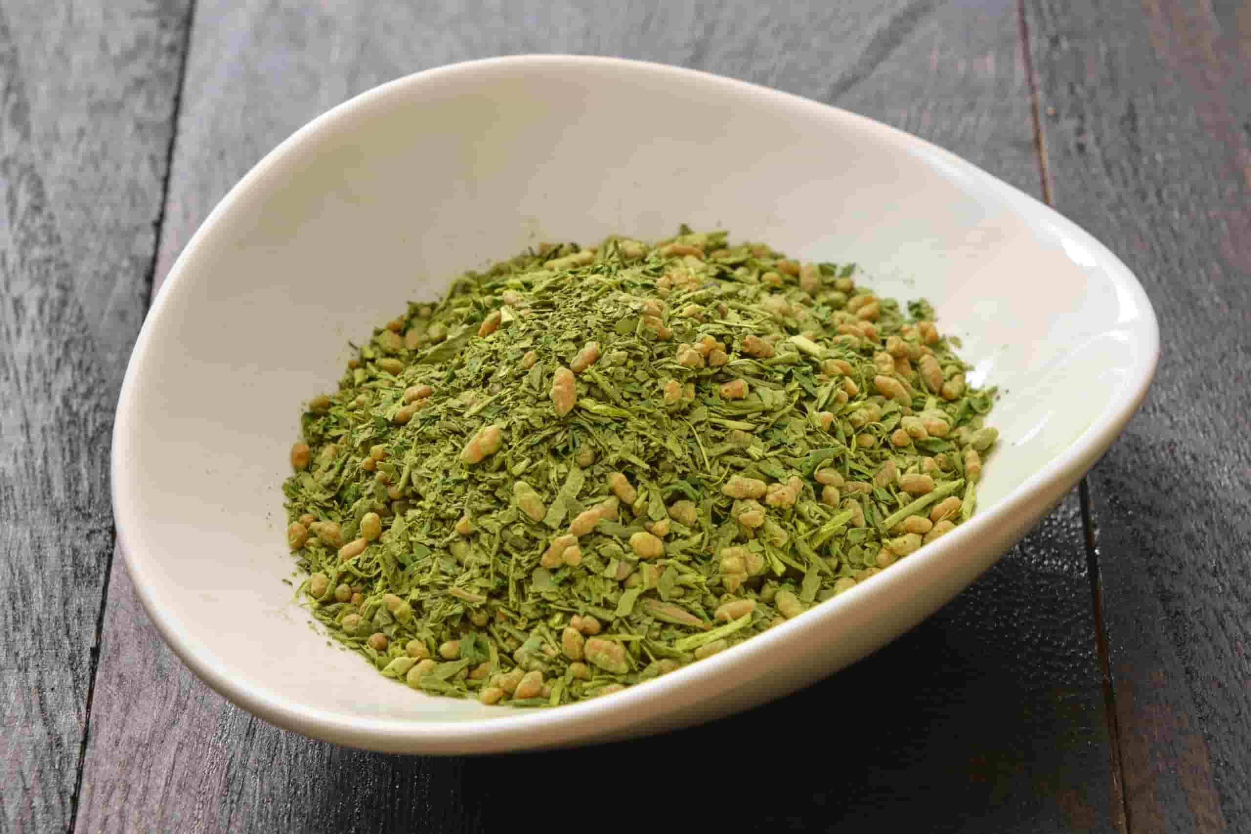 A white bowl filled with green genmaicha tea leaves blended with roasted brown rice, placed on a dark wooden table.