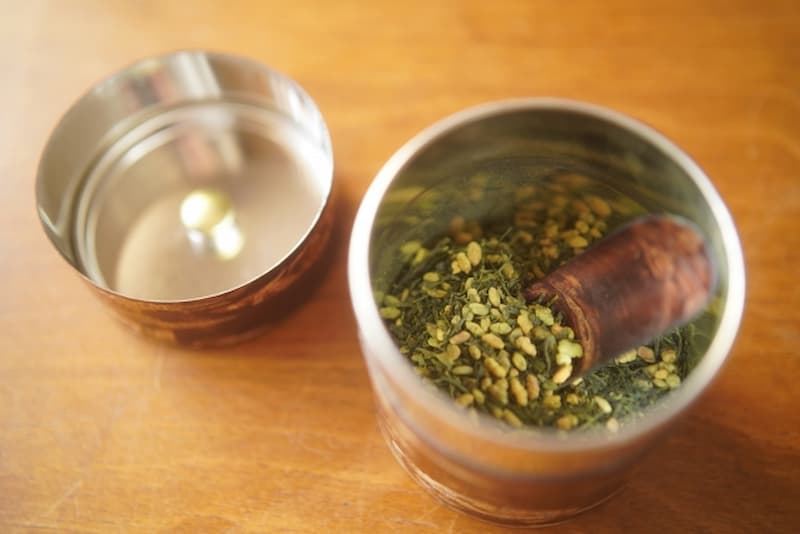 A bowl of matcha-iri genmaicha containing green tea leaves and roasted rice, with a wooden scoop resting inside, beside an empty metal container on a wooden table.