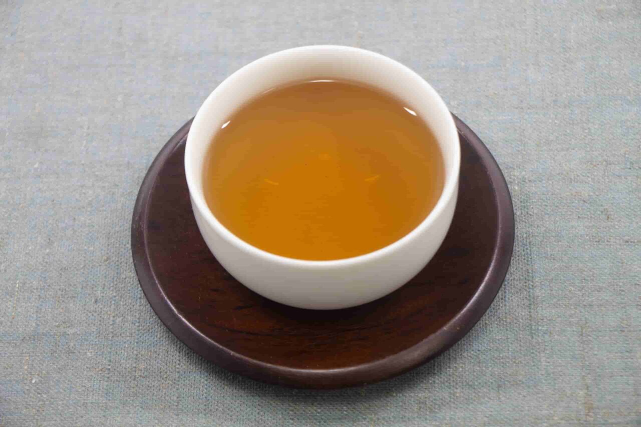 A cup of golden-brown sannenbancha tea served in a white cup on a dark wooden saucer.