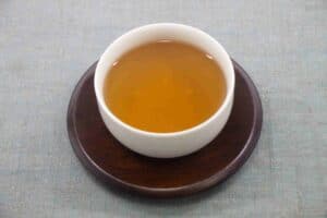 A cup of golden-brown sannenbancha tea served in a white cup on a dark wooden saucer.
