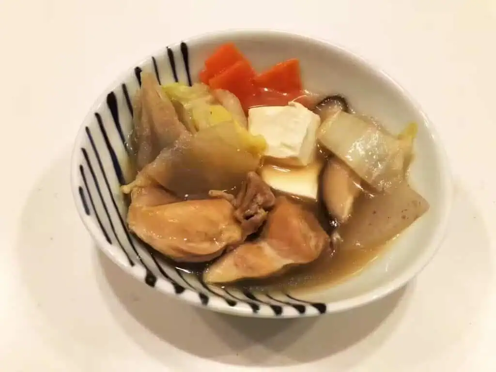 A bowl of iriyaki with chicken, tofu, napa cabbage, carrot and konnyaku in a blue-striped white bowl