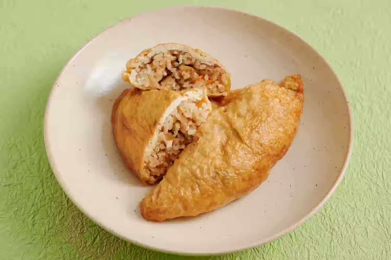 Two triangular itadaki, rice-stuffed fried tofu pouches, on a white plate against a green paper background
