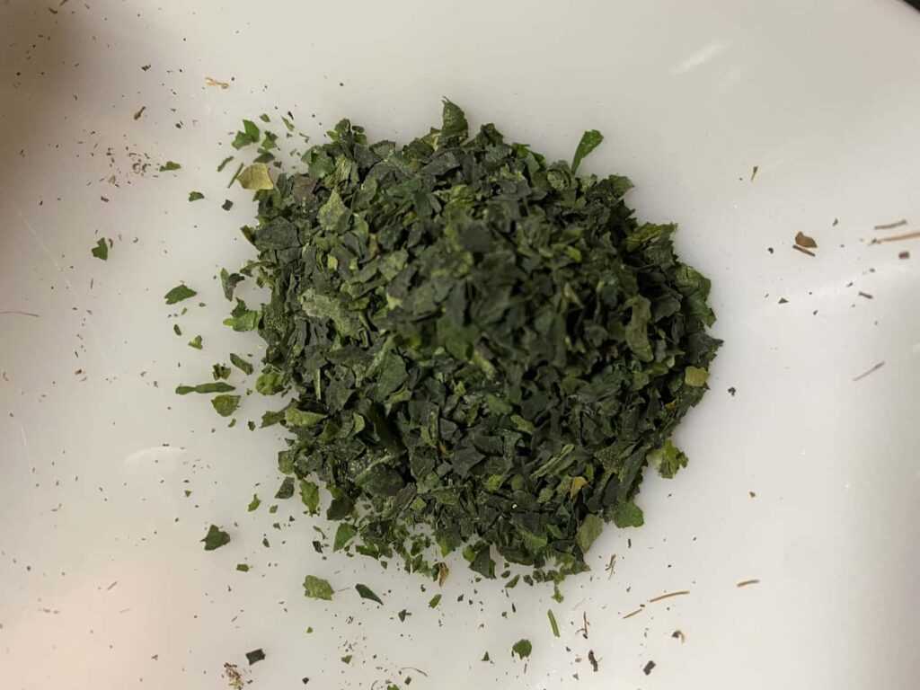 A pile of dark green tencha tea leaves, roughly chopped into small flakes, on a white ceramic plate.
