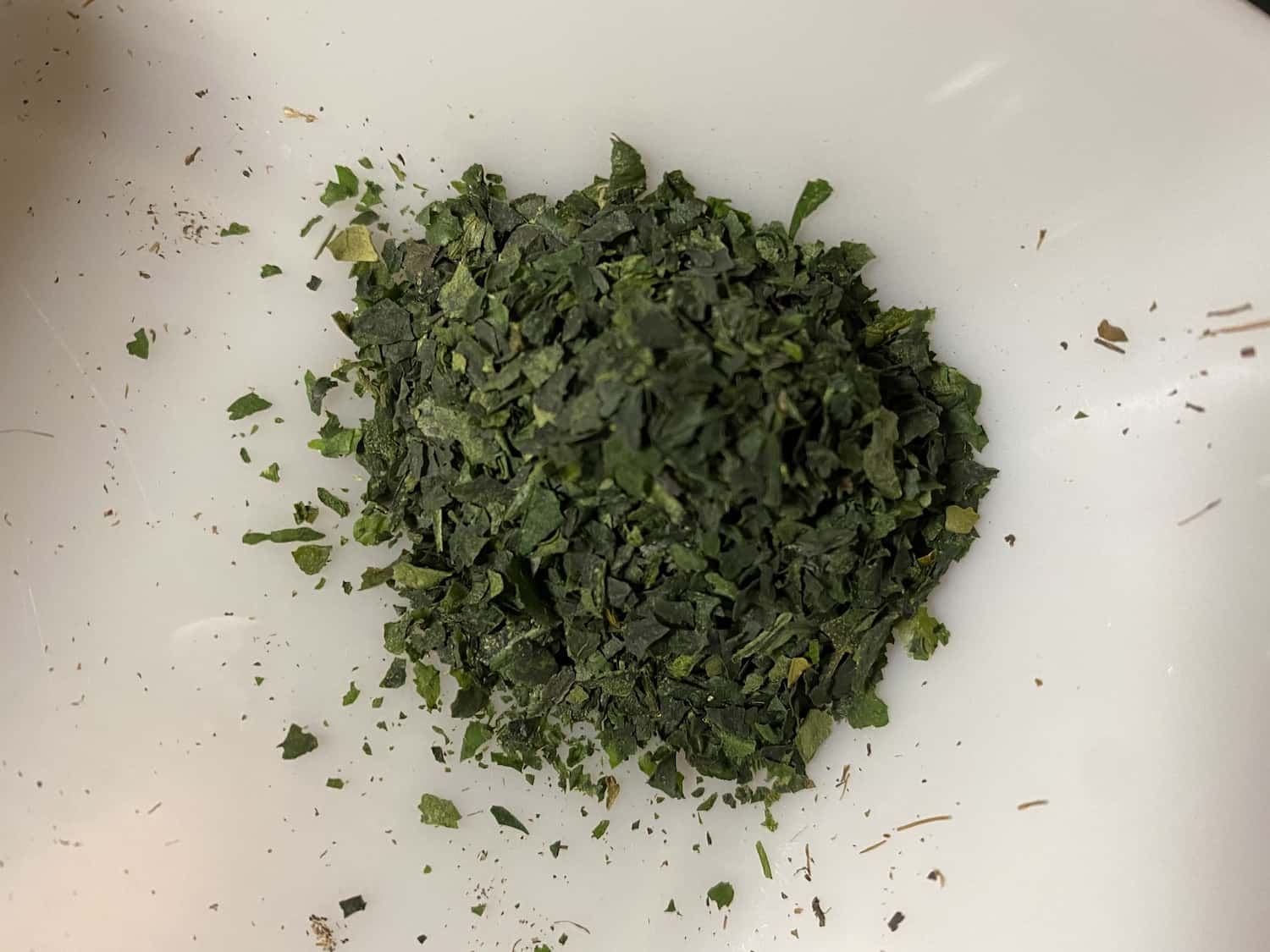 A pile of dark green tencha tea leaves, roughly chopped into small flakes, on a white ceramic plate.
