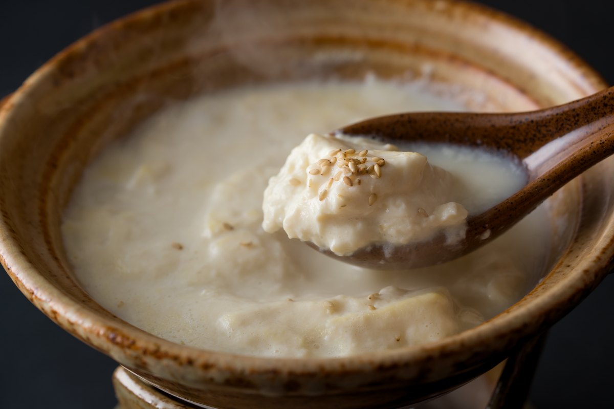 A spoon lifting soft tofu with sesame seeds from a clay pot of milky Ureshino Onsen Yudofu broth